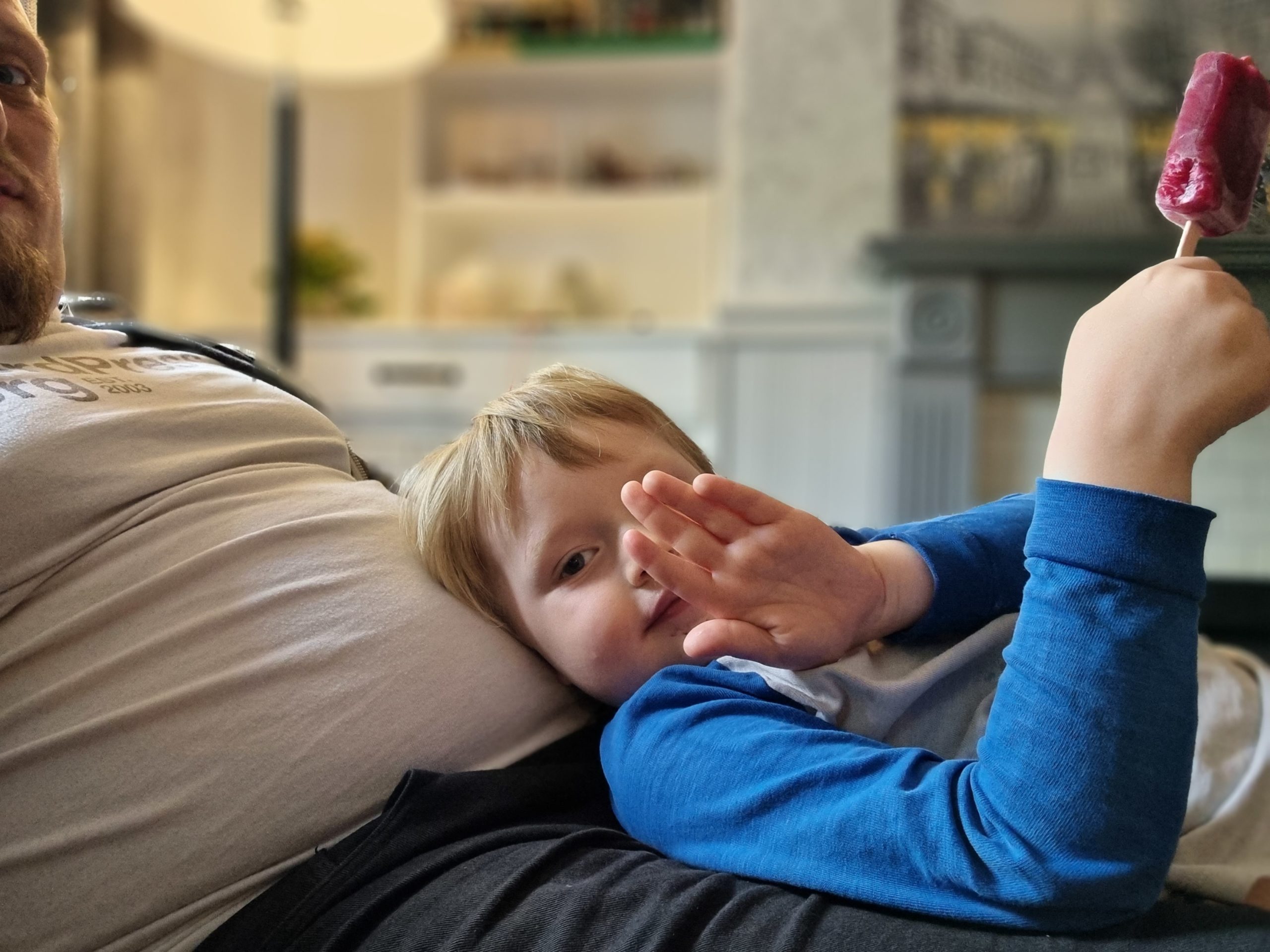 Full image of Boy lying on Dan, waving, eating an ice lolly.