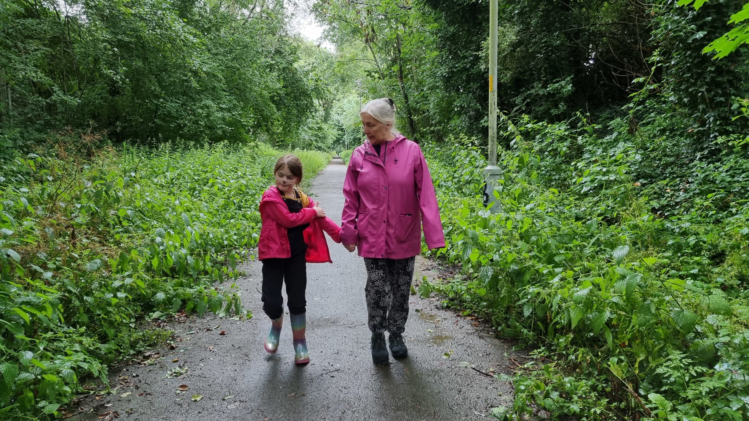 Full image of A woman and a girl on a cycleway, holding hands, between trees and weeds.