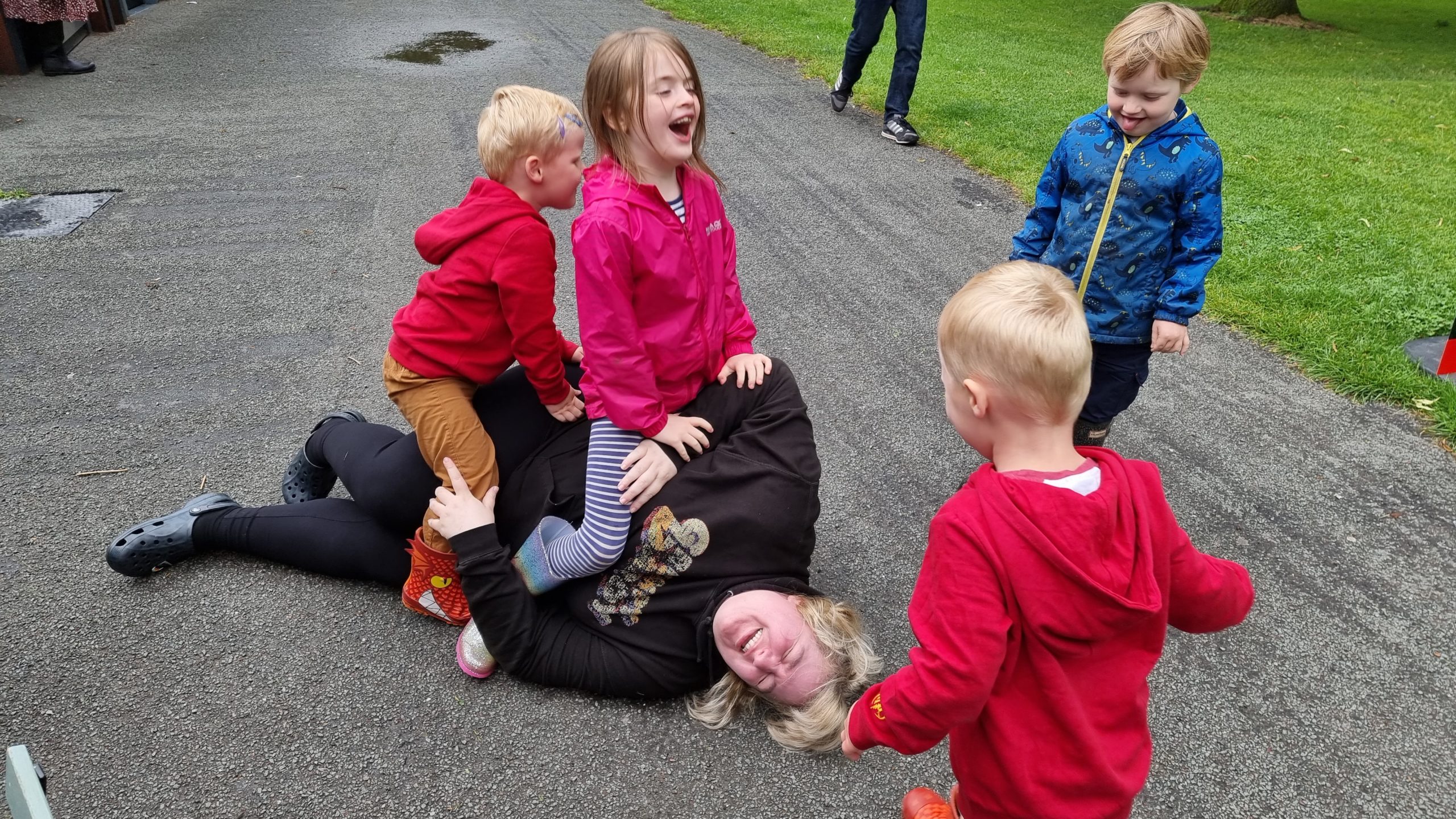 Full image of Becky lies on the floor, surrounded by/covered in children.