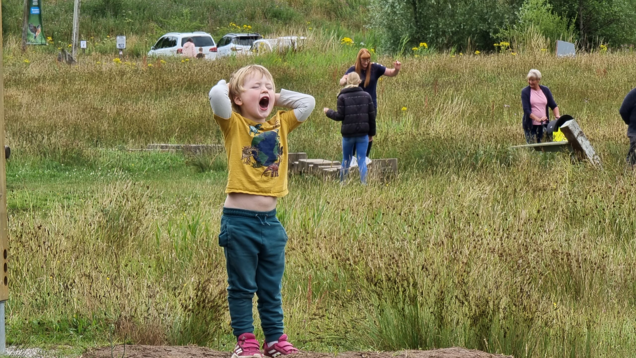Full image of A boy shouts from the top of a grassy knoll near a car park.