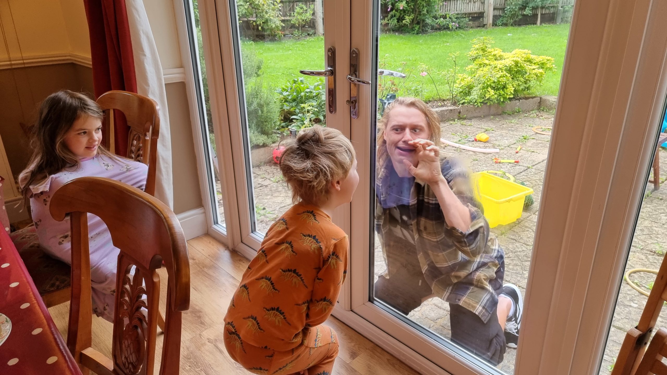 Full image of Children in pyjamas at a breakfast table greet Robin outside the patio doors.