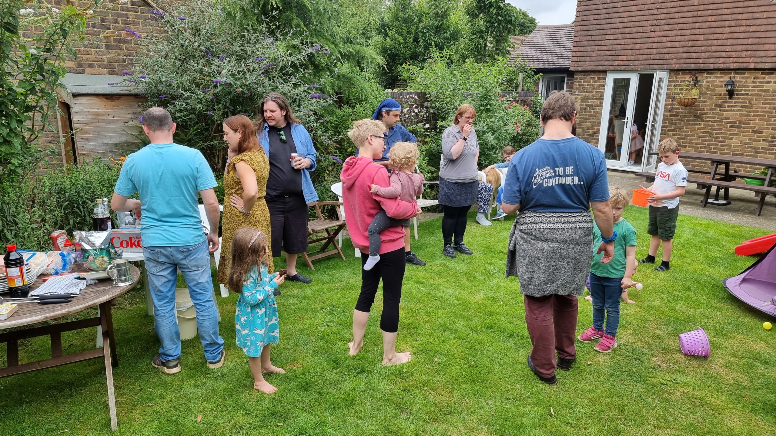 Full image of People stand around in a garden at a barbecue.