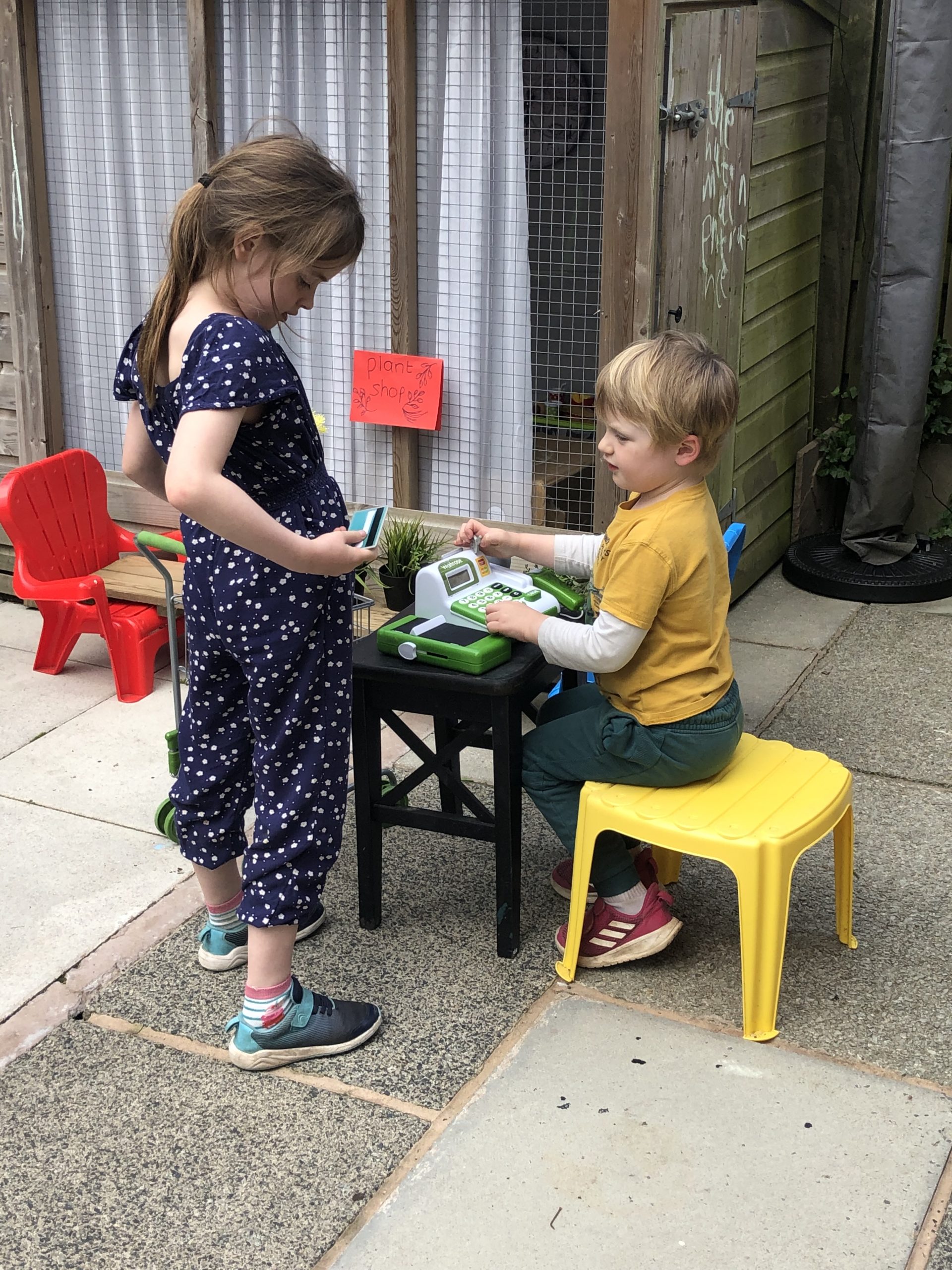 Full image of Children play shop: a boy operates a checkout and a girl buys shopping.