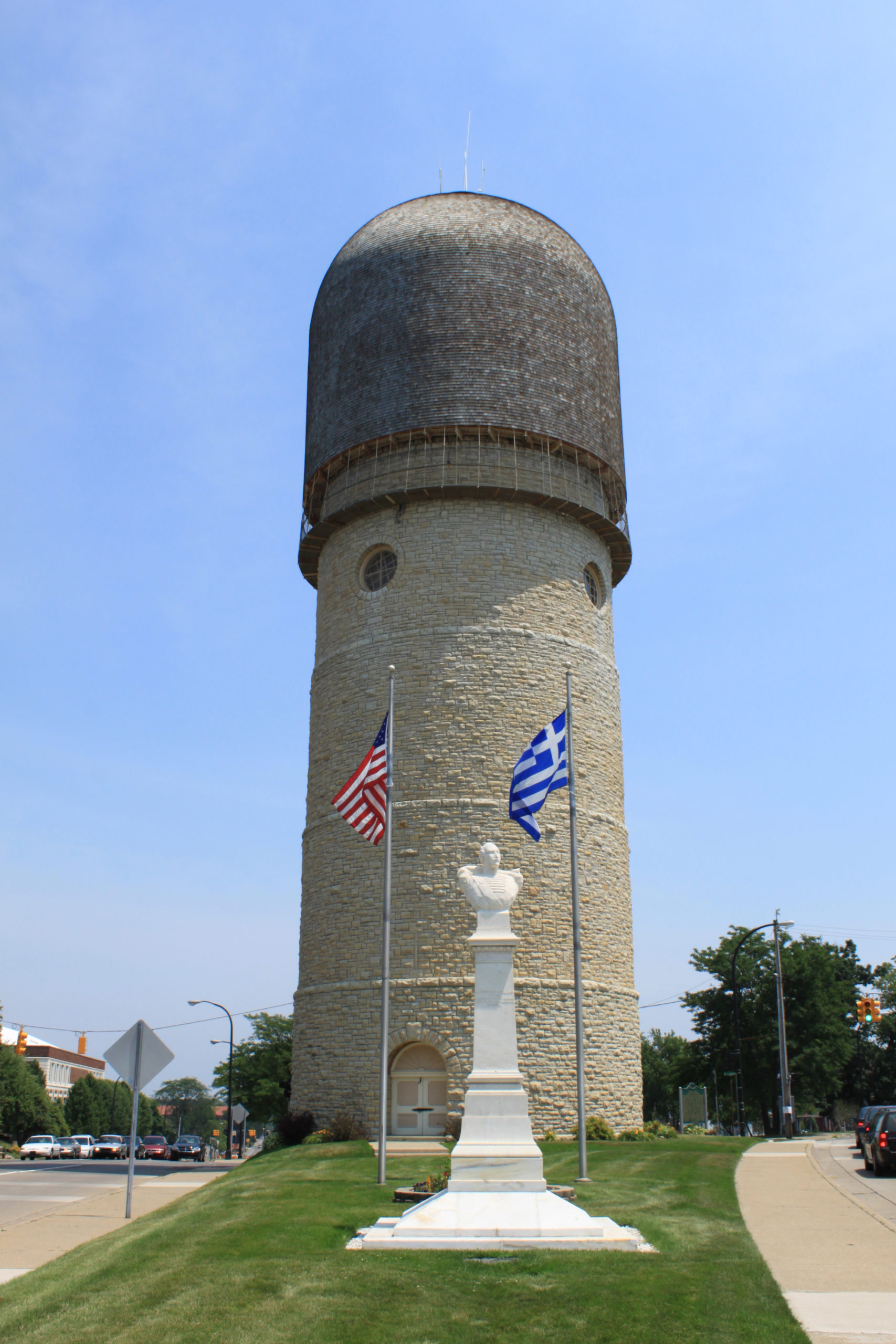 Full image of The Ypsilanti Water Tower, at the intersection of Washtenaw Avenue and Cross Street, Ypsilanti, Michigan. The tower is listed in the National Register of Historic Places, and is a National Historic Civil Engineering Landmark. An American flag and a Greek flag are flying, and a bust of the Greek general, Demetrios Ypsilantis (also commonly spelled "Demetrius Ypsilanti"), for whom the city is named, is in the foreground. Photo by Dwight Burdette, used under a Creative Commons license.