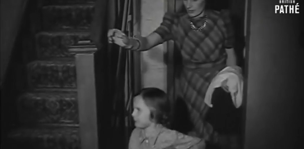 Black & white framegrab showing a woman following her child, wearing pyjamas, towards a staircase up.