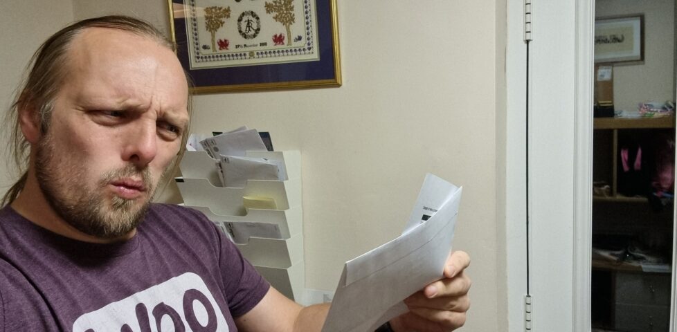Dan, near his front door, reads his mail. His facial expression suggests that he's about to exclaim "What!?"