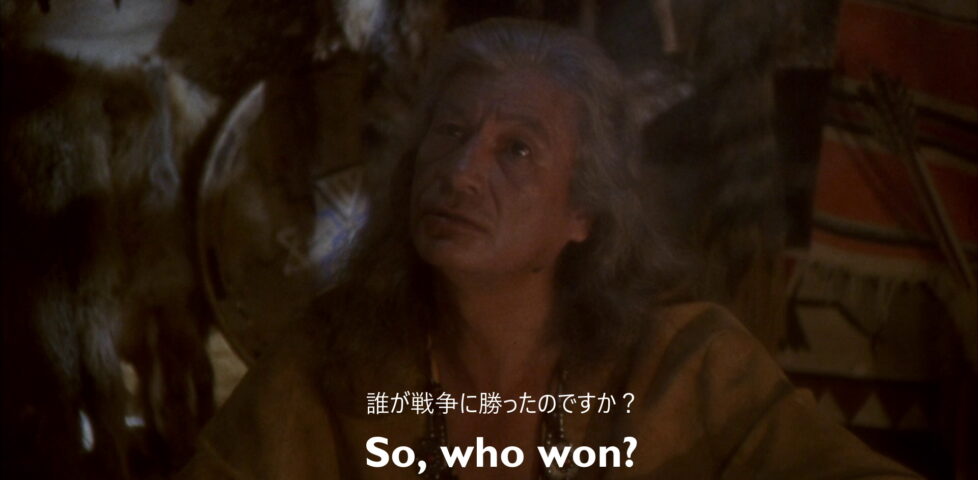Still from Hot Shots!. Owatonna, an older Native American man, sits surrounded by animal skins. An English subtitle reads "So, who won?" A Japanese subtitle reads "誰が戦争に勝ったのですか?", which translates as "Who won the war?"
