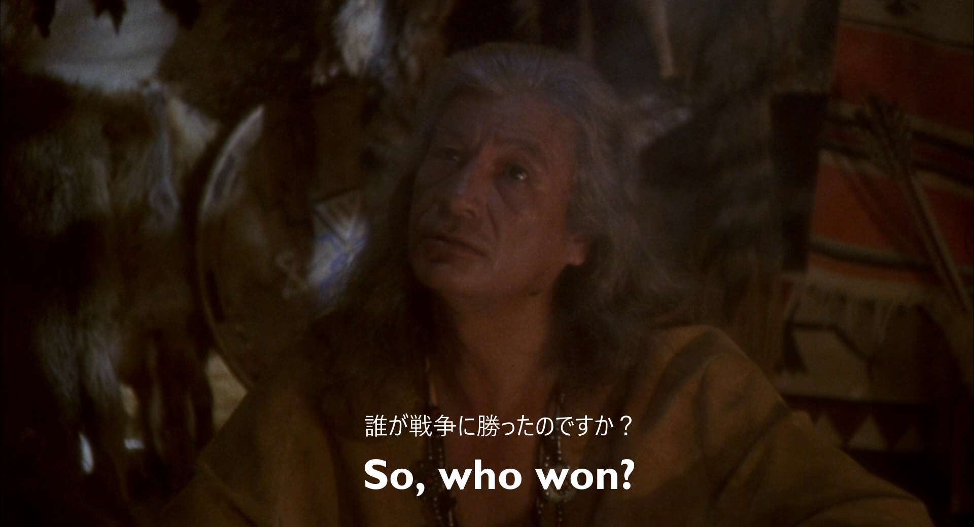 Full image of Still from Hot Shots!. Owatonna, an older Native American man, sits surrounded by animal skins. An English subtitle reads "So, who won?" A Japanese subtitle reads "誰が戦争に勝ったのですか?", which translates as "Who won the war?"