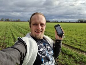 Dan in a grassy field holding a GPSr and grinning