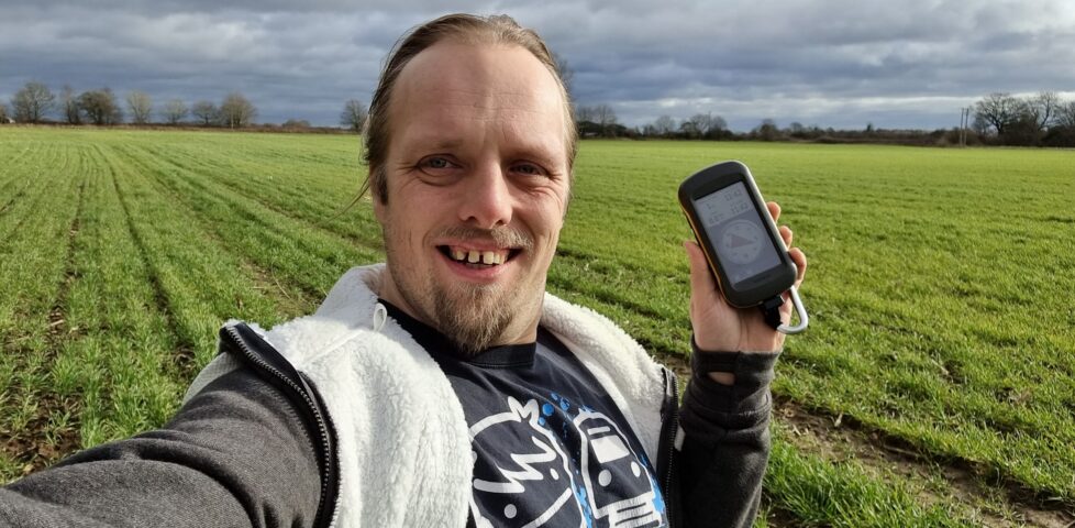 Dan in a grassy field holding a GPSr and grinning