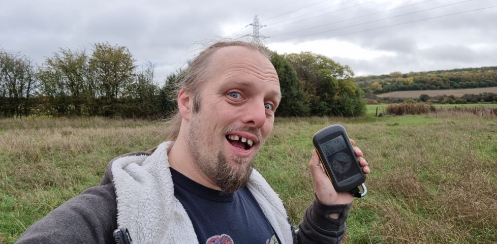 Dan makes a silly grin while holding a GPS receiver in a damp grassy autumn meadow, on a grey and drizzly day.