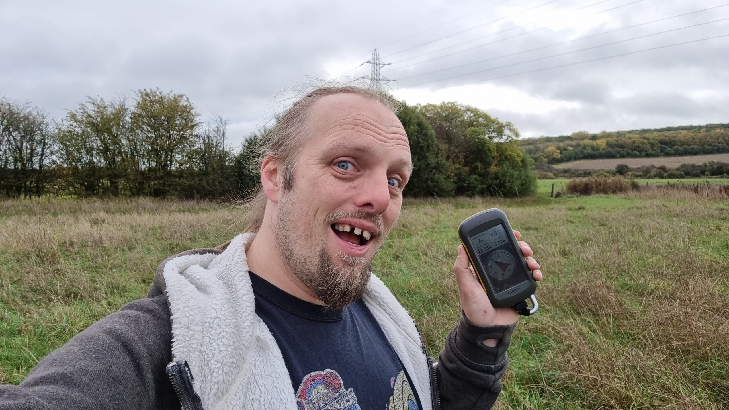Full image of Dan makes a silly grin while holding a GPS receiver in a damp grassy autumn meadow, on a grey and drizzly day.