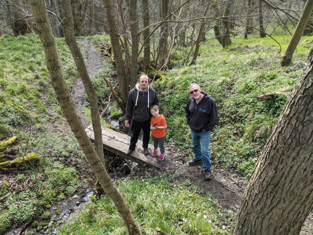 Dan and John stand on a two-plank footbridge over a stream alongside Dan's fellow Three Rings volunteer Paul, looking uphill towards the camera. The stream runs through moderately-dense deciduous woodland and the forest floor is covered with greenery.