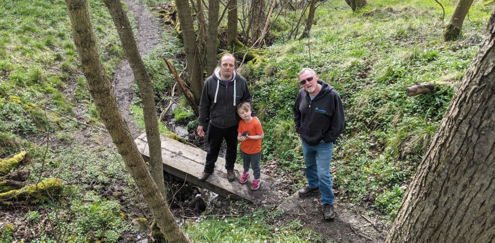 Dan and John stand on a two-plank footbridge over a stream alongside Dan's fellow Three Rings volunteer Paul, looking uphill towards the camera. The stream runs through moderately-dense deciduous woodland and the forest floor is covered with greenery.