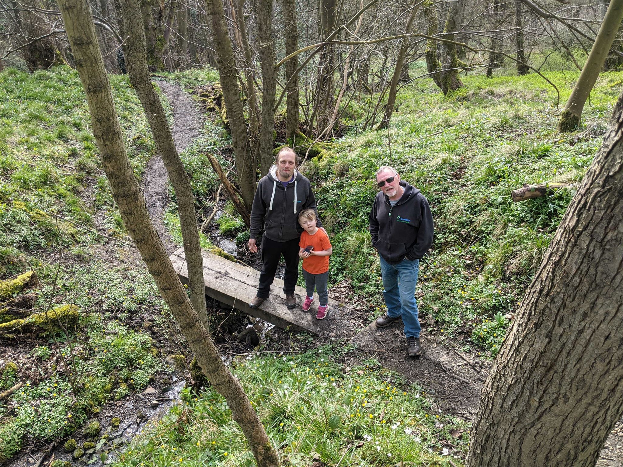 Full image of Dan and John stand on a two-plank footbridge over a stream alongside Dan's fellow Three Rings volunteer Paul, looking uphill towards the camera. The stream runs through moderately-dense deciduous woodland and the forest floor is covered with greenery.