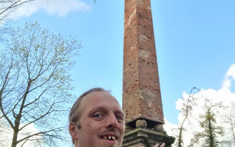 Dan stands under a brick obelisk under a mostly-clear blue sky. He is smiling and pointing at the landmark.