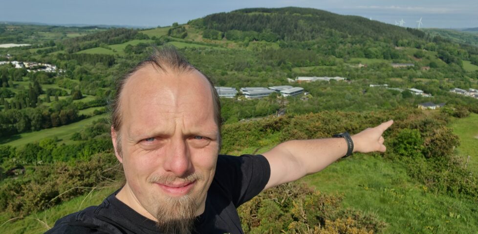 Dan, on a hilltop, points down into the valley between this and the next hill over. Where he's pointing is a white-and-brown-brick building with a grey slate roof in the iconic shape of a Travelodge hotel.