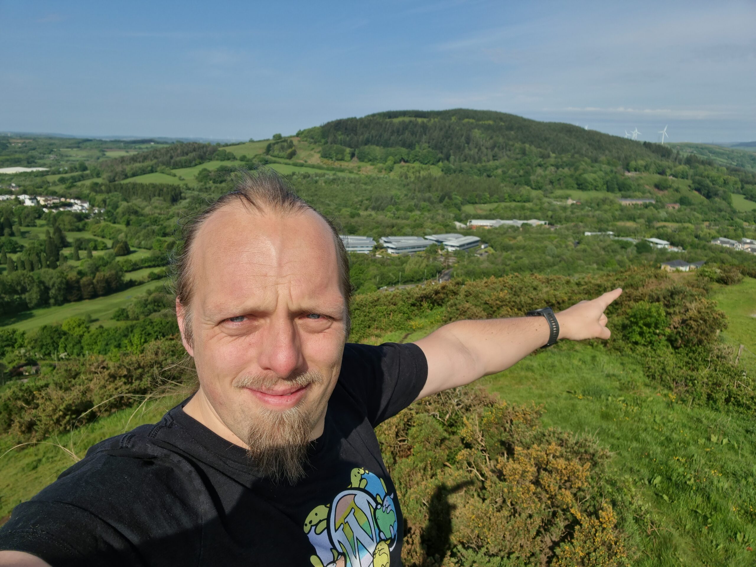 Full image of Dan, on a hilltop, points down into the valley between this and the next hill over. Where he's pointing is a white-and-brown-brick building with a grey slate roof in the iconic shape of a Travelodge hotel.