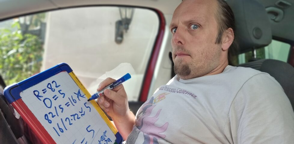 Dan sitting in the driving seat of a car, doing maths on a portable whiteboard.