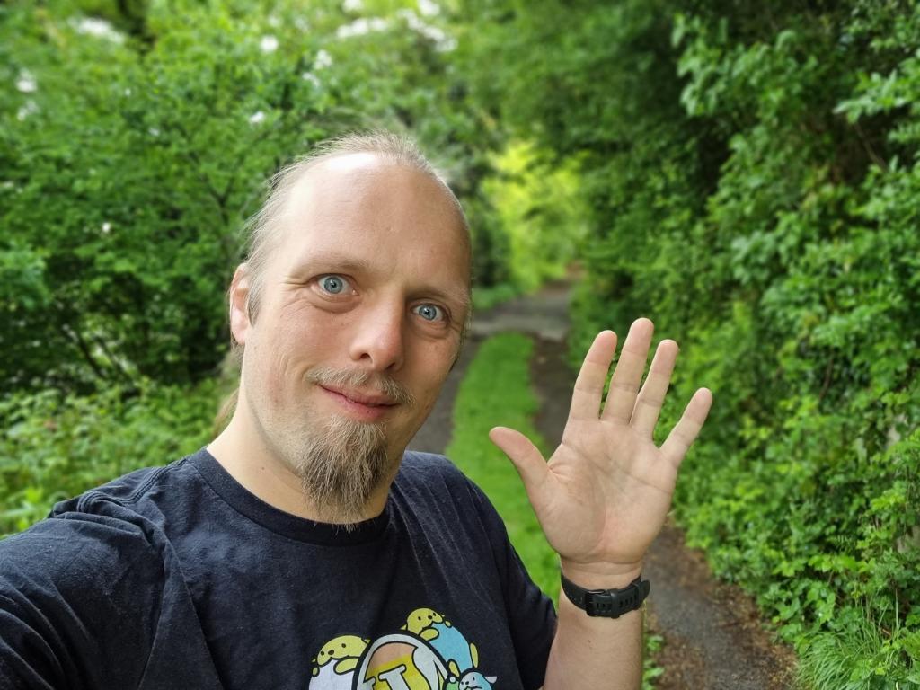 Dan stands on a rough muddy forest trail ad waves at the camera.