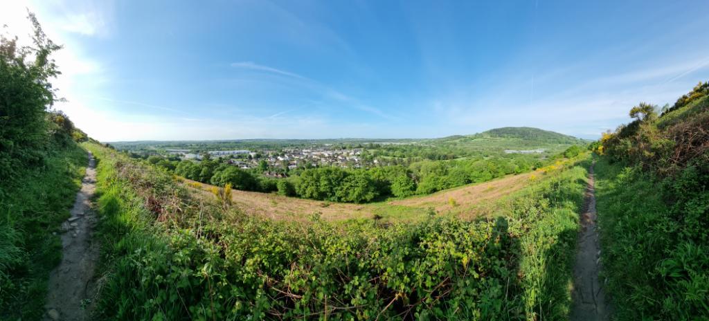 Panoramic view showing a small settlement nestled between rolling hills. The skies are clear blue streaked by long wispy clouds, and a narrow footpath runs from right to left between thorny bushes on a grassy hillside.