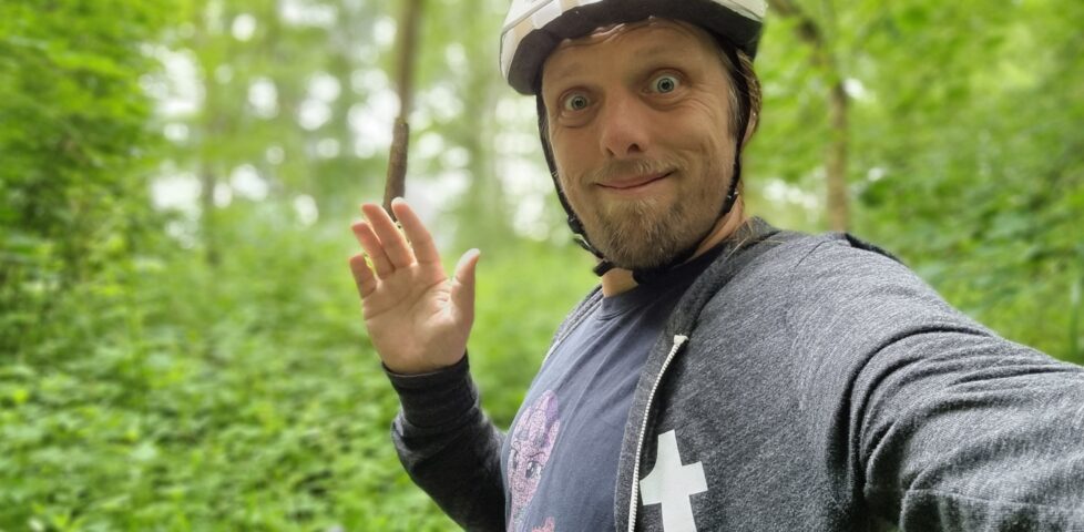 Dan, wearing a Tumblr hoodie over a My Little Pony T-shirt and with a white cycle helmet on his head, stands in front of his bike (with a GPS receiver on the handlebars) in a deep green forest.