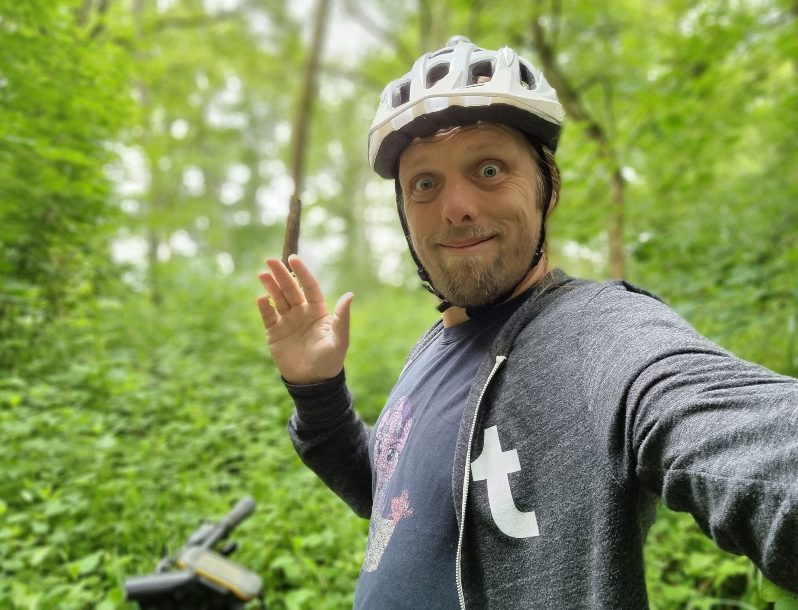 Full image of Dan, wearing a Tumblr hoodie over a My Little Pony T-shirt and with a white cycle helmet on his head, stands in front of his bike (with a GPS receiver on the handlebars) in a deep green forest.