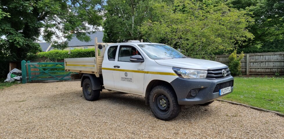 A rugged white flatbed van with a yellow stripe and the logo of Oxfordshire County Council sits on a gravel driveway in a garden. The sun glistens off the windscreen and the driver-side window is partially ajar.