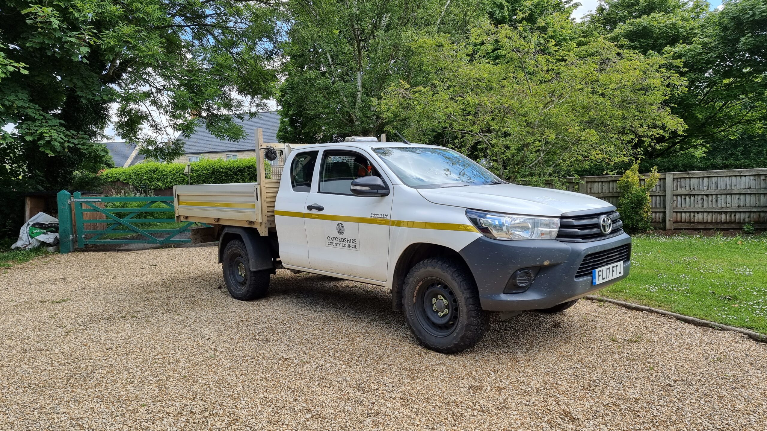 Full image of A rugged white flatbed van with a yellow stripe and the logo of Oxfordshire County Council sits on a gravel driveway in a garden. The sun glistens off the windscreen and the driver-side window is partially ajar.