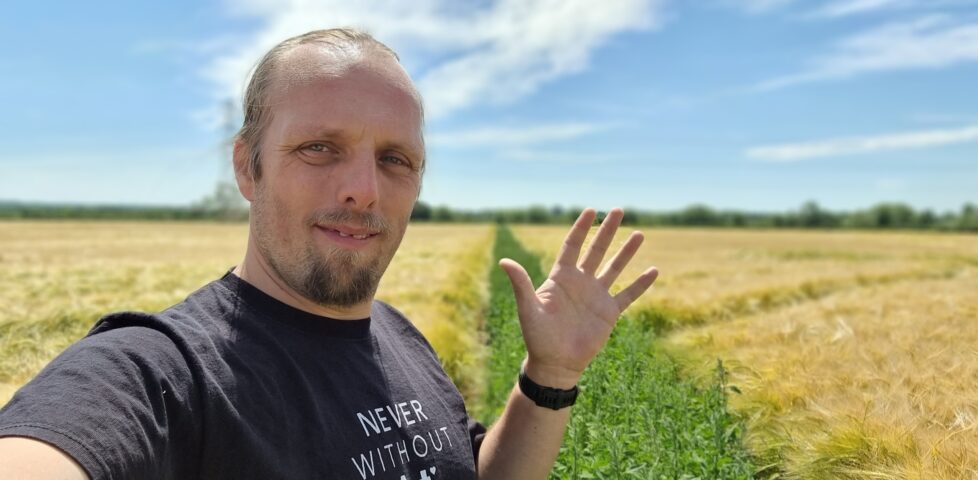 Dan, in a black t-shirt with "never without art" written on it, waves to the camera. He's standing in a cornfield, but the "path" that cuts diagonally through it is filled with plants to a similar height as the corn, making it hard to pass.