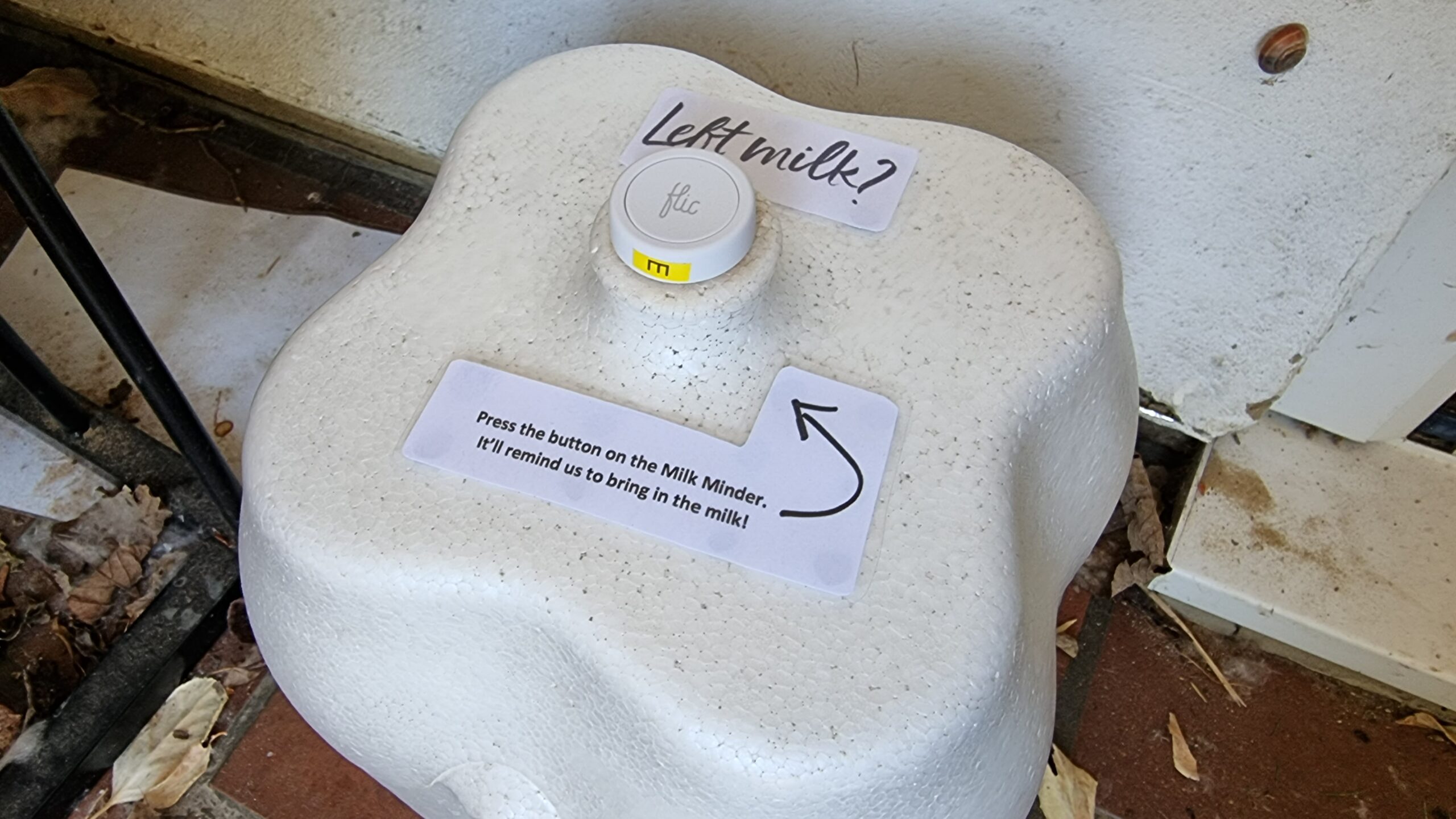 Full image of Milk container, with a Flic 2 button attached to the handle of the lid and a laminated notice attached, reading: "Left milk? Press the button on the Milk Minder. It'll remind us to bring in the milk!"