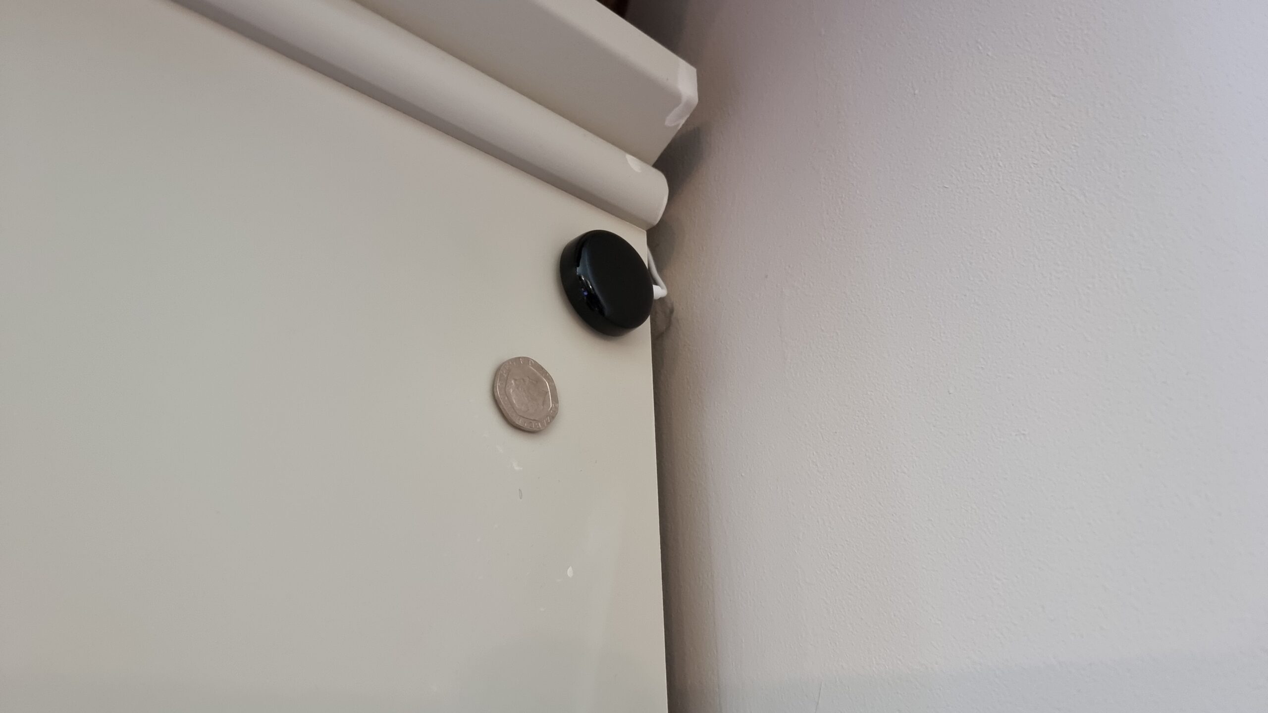 Full image of Flic Infared Transceiver on the side of a bookcase, alongside an (only slighter smaller than it) 20p piece, for scale.