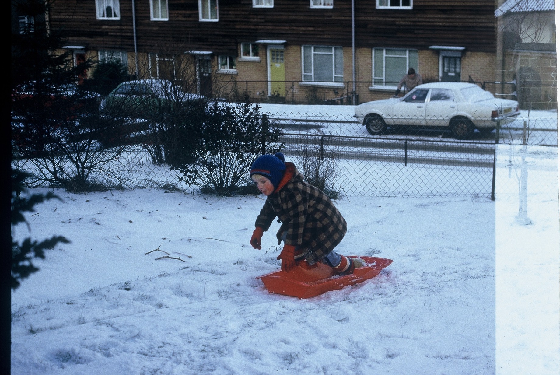 Full image of Dan, aged around 4, dressed in a duffel coat, bobble hat and gloves, kneeling on a red plastic sledge in a snow-filled garden. The garden is bordered by a wire fence, and in the background a man can be seen scraping icee off a car.