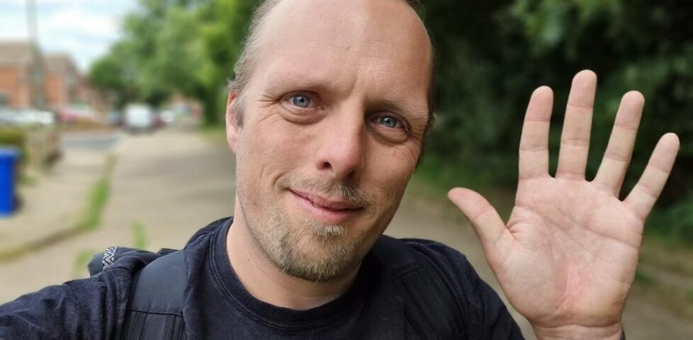Dan, in a black t-shirt, waves to the camera from a suburban street.