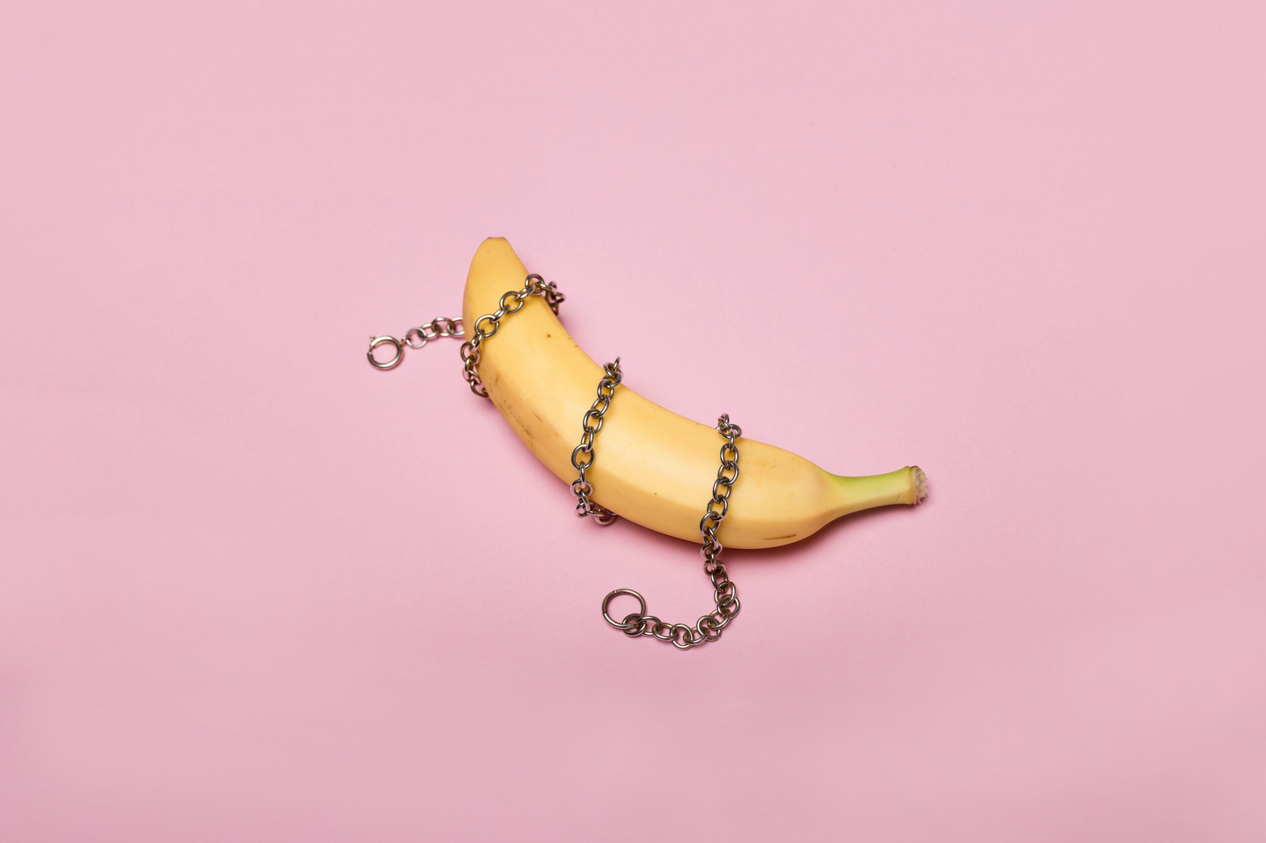 Full image of Photograph showing a yellow banana on a pink background. The banana has a silver chain wrapped around it three times. Photo courtesy Deon Black.