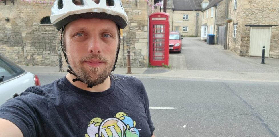 Dan, in a cycle helment, stands on a street corner. A red telephone box is visible in the background.