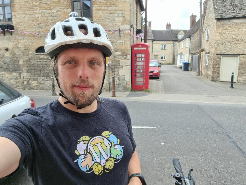 Dan, in a cycle helment, stands on a street corner. A red telephone box is visible in the background.