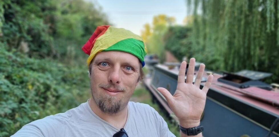 Dan, wearing a rainbow-striped bandan, waves to the camera from a rural canal towpath.