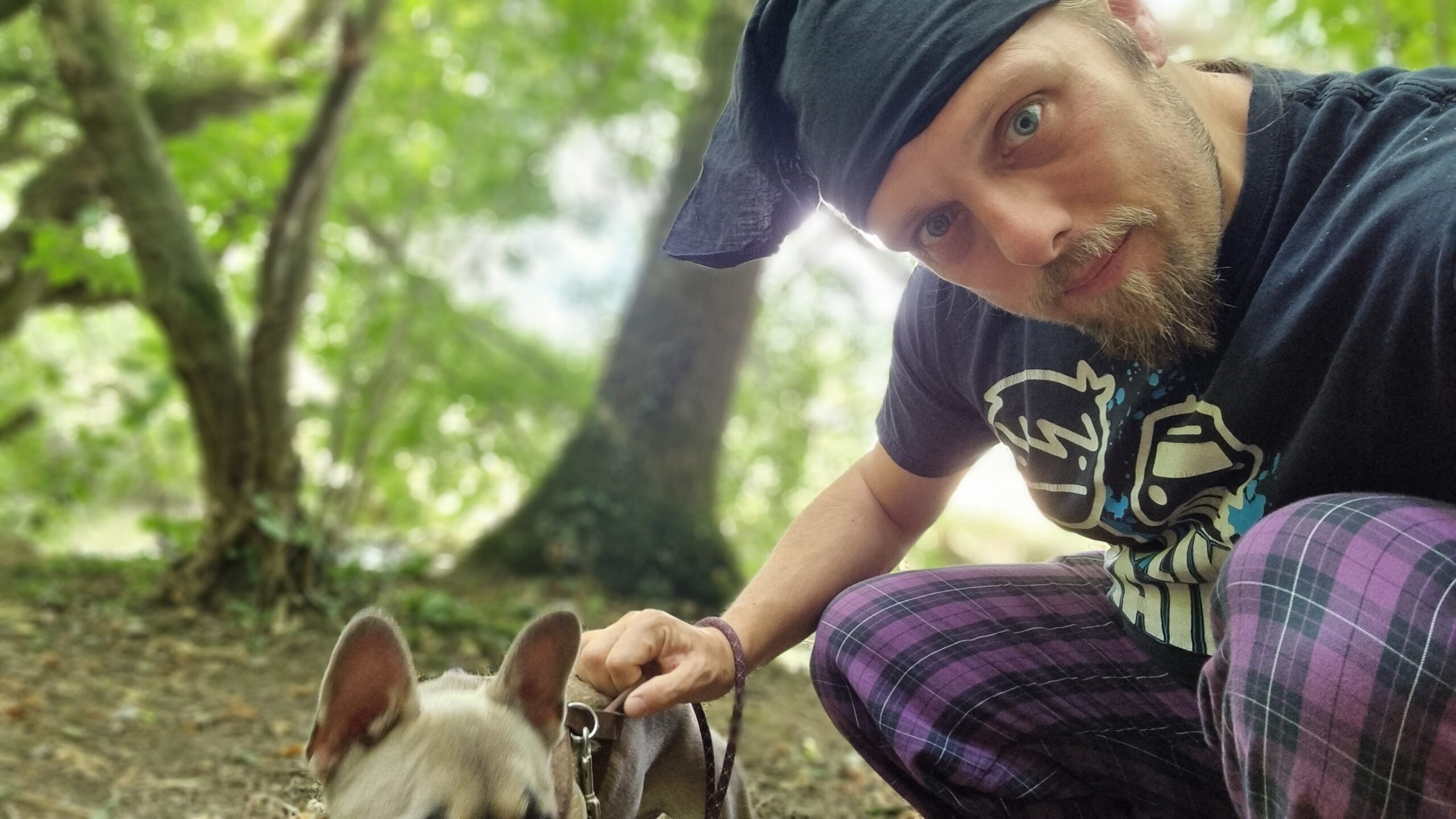 Full image of Dan, wearing purple pyjama bottoms, a black t-shirt, and a black bandana, sits in a woodland setting with a dog who's just put herself off the bottom of the frame at the last second.