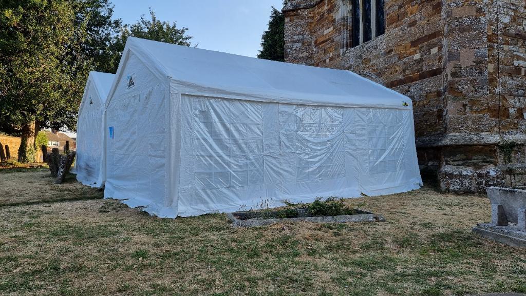 An enclosed tempory gazebo with a tentlike covering, at the foot of a stone church tower.