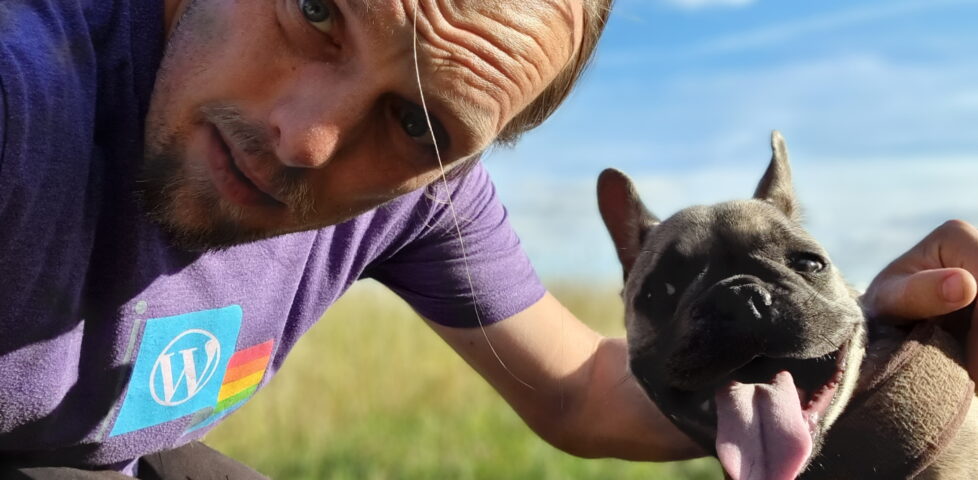 Under bright blue skies and in a green field, Dan and Demmy stare into the camera. Dan's flushed from the heat and wearing a purple-and-rainbow "WordPress Pride" t-shirt; Demmy's tongue hangs low and she's clearly panting.