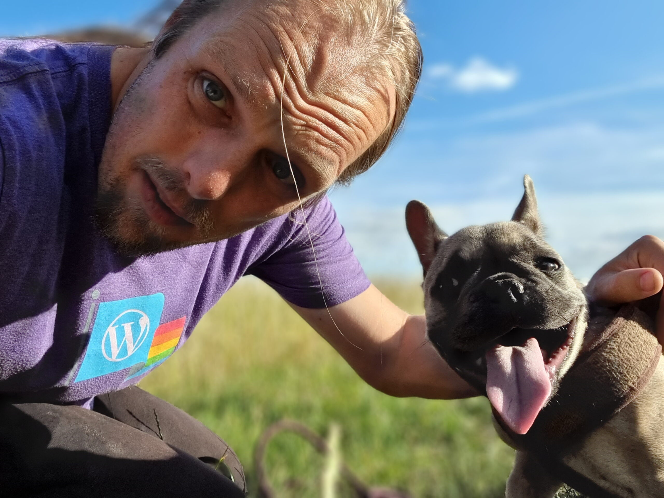 Full image of Under bright blue skies and in a green field, Dan and Demmy stare into the camera. Dan's flushed from the heat and wearing a purple-and-rainbow "WordPress Pride" t-shirt; Demmy's tongue hangs low and she's clearly panting.