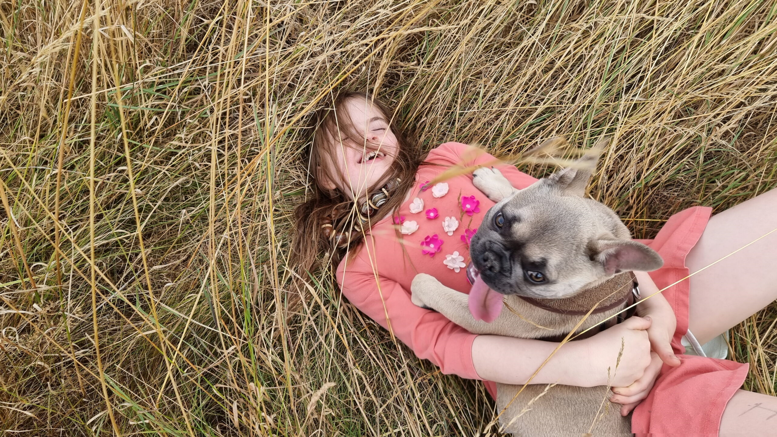 Full image of An 8-year-old girl in a flowery pink dress lies on her back in a meadow of long dry grass, laughing with her eyes closed. On her belly, Demmy lies, looking up towards the camera with her tongue hanging out.