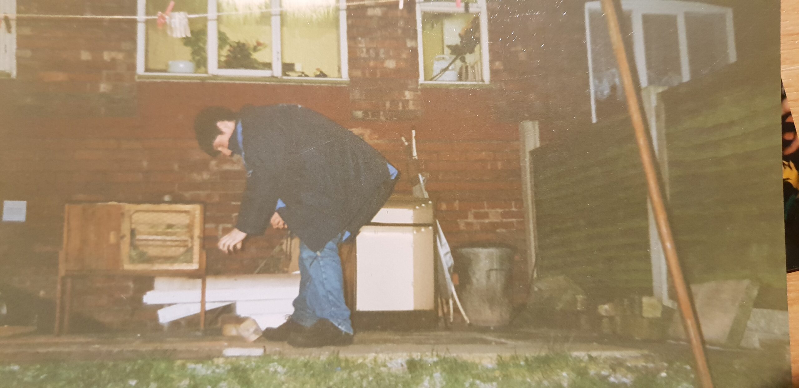 Full image of A young man in jeans and a blue coat stands on the patio in the back garden of a terraced house, dropping a half-brick onto the floor. In the background, an unused rabbit hutch and a dustbin can be seen. The photo is clearly taken using a flash, at night.