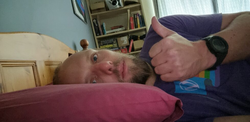 Dan lying in bed, giving a weak "thumbs up".