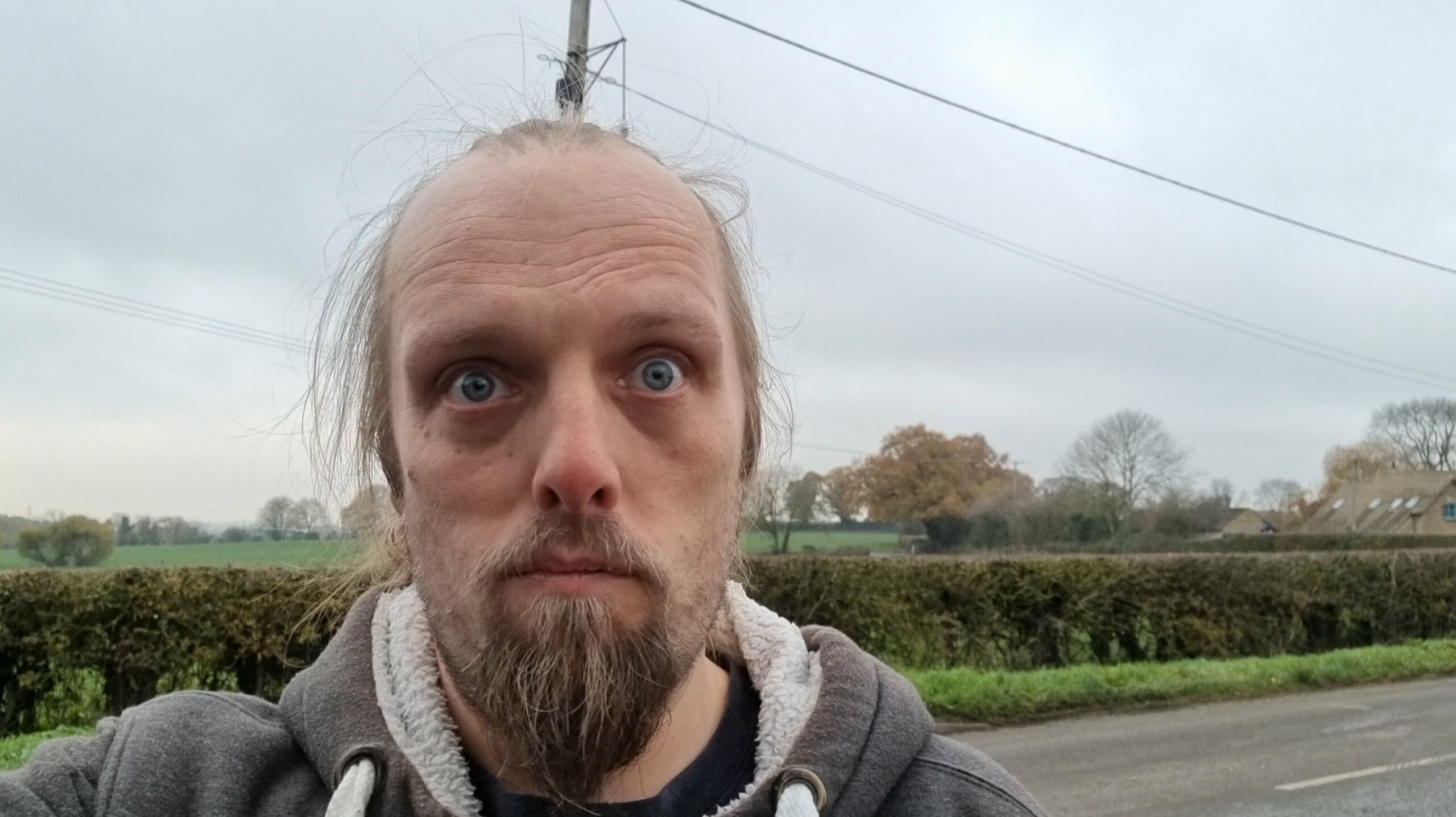 Full image of Standing in front of an advancing cloud front in a rural village, Dan - dressed in a grey and white Three Rings hoodie - stares sadly into the camera.
