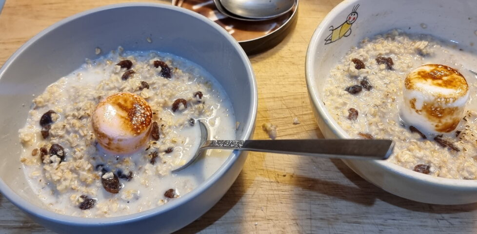 Two bowls of porridge with sultanas, each topped with a jumbo marshmallow (one pink, one white) being toasted by a blowtorch (mostly off-camera).