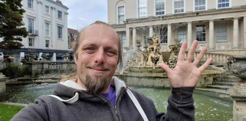 In an urban environment, Dan waves. Behind him, a stone fountain-statue representing Neptune, surrounded by sea creatures, can be seen.