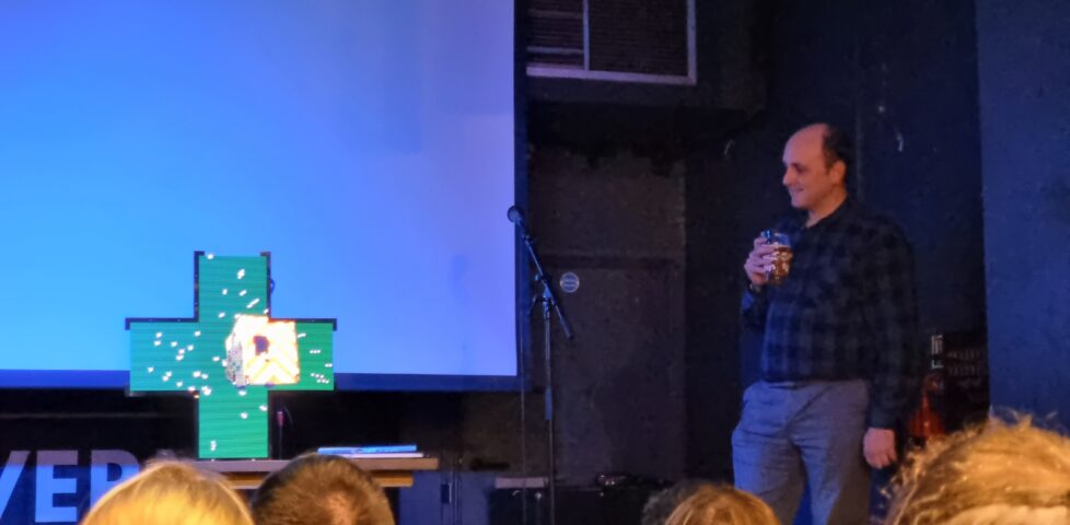 Matt Westcott stands to the side of a stage, drinking beer, while centrestage a cross-shaped "pharmacy sign" projects an animation of an ambulance rocketing into a starfield.