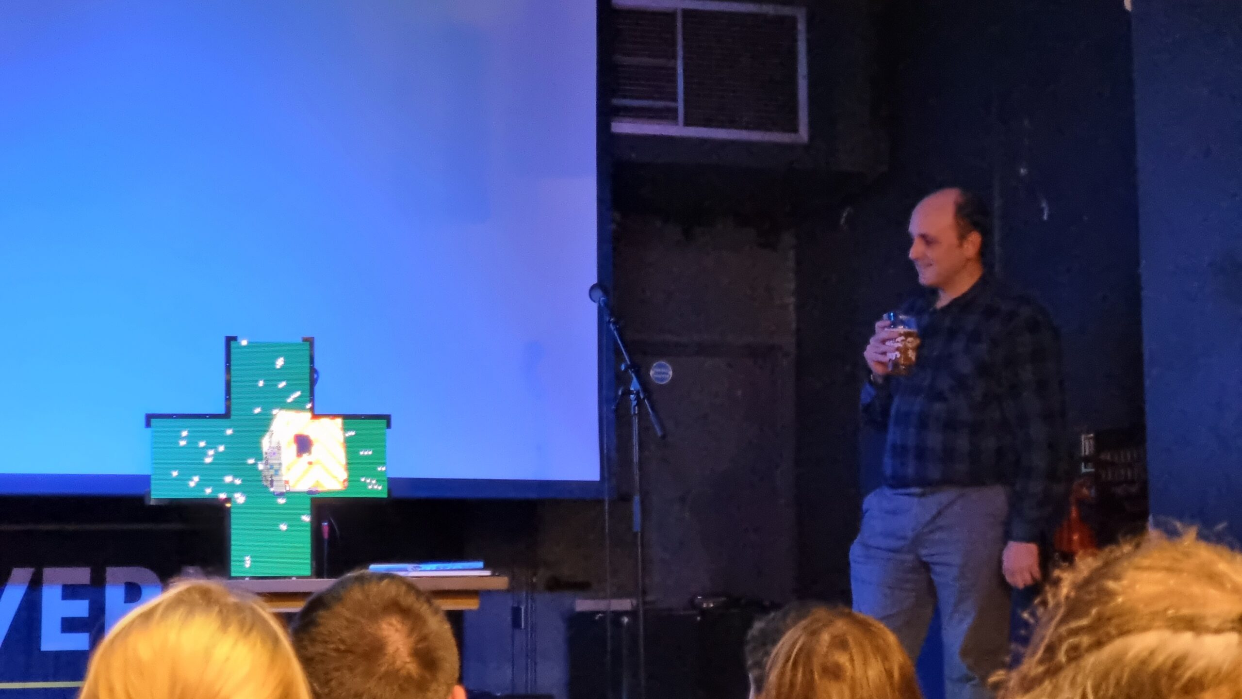 Full image of Matt Westcott stands to the side of a stage, drinking beer, while centrestage a cross-shaped "pharmacy sign" projects an animation of an ambulance rocketing into a starfield.